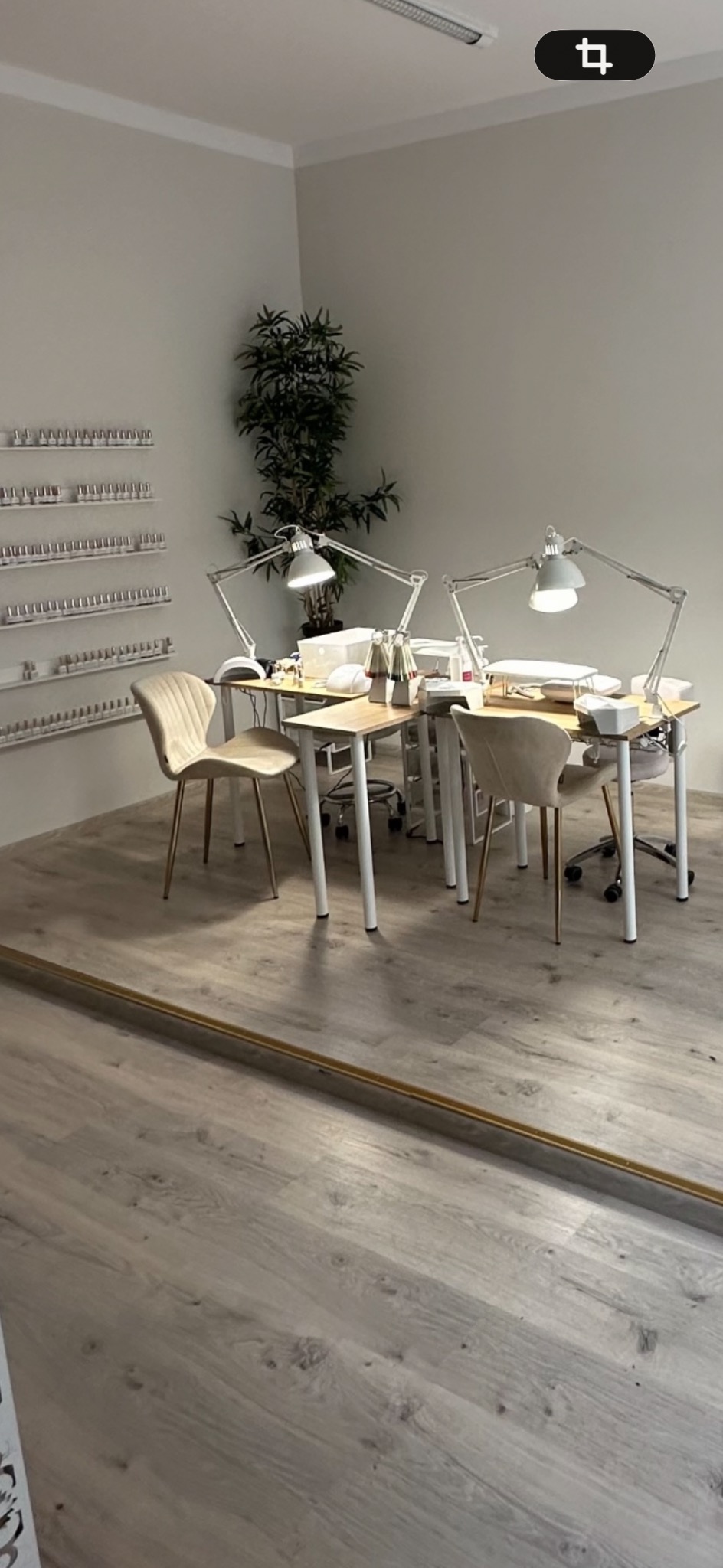 Jasne wnętrze salonu kosmetycznego z dwoma stanowiskami do manicure, wyposażonymi w lampy i akcesoria. Na ścianie półki z lakierami, w rogu zielona roślina.