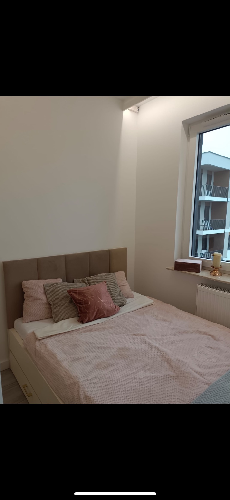 Jasna sypialnia z beżowym zagłówkiem łóżka, różową pościelą i dekoracyjnymi poduszkami. Za oknem widoczny budynek z balkonami. Minimalistyczny wystrój.