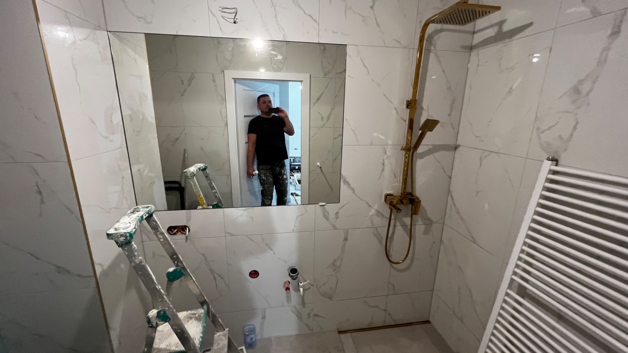 Wykończona łazienka z białymi płytkami imitującymi marmur, złotą armaturą i drabinkowym grzejnikiem. W lustrze odbija się osoba i drabina malarska.