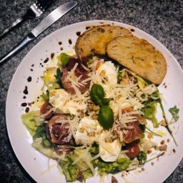 Przy Stole Catering - Sałatka z mozzarellą, szynką parmeńską, pomidorkami cherry, grzankami i sosem balsamicznym na białym talerzu, obok nóż i widelec.