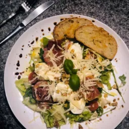 Sałatka z mozzarellą, szynką parmeńską, pomidorkami cherry, grzankami i sosem balsamicznym na białym talerzu, obok nóż i widelec.