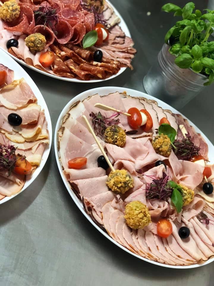 Trzy białe półmiski z różnymi rodzajami wędlin, serów i przekąsek, udekorowane pomidorkami cherry, oliwkami i ziołami, obok doniczka z bazylią.