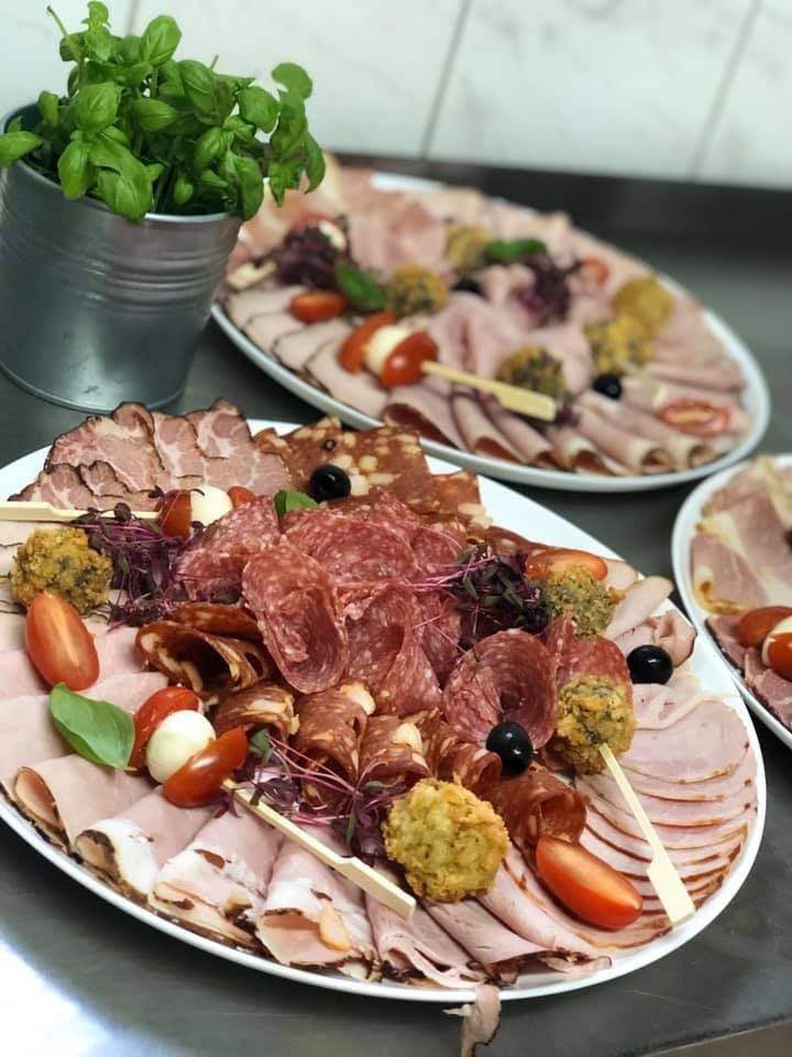 Dekoracyjnie ułożone wędliny, sery i warzywa na białych półmiskach, z doniczką bazylii w metalowym wiaderku w tle.