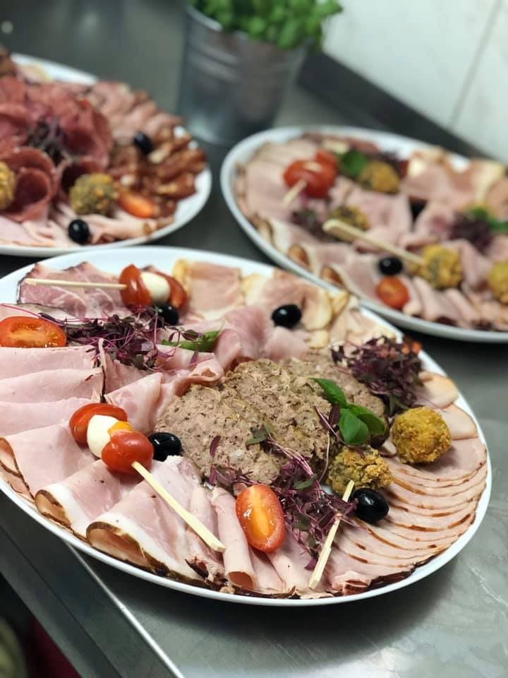 Trzy białe talerze z różnorodnymi wędlinami, pastami i przekąskami, udekorowane pomidorkami cherry, oliwkami i świeżymi ziołami, ustawione na metalowym blacie w tle widoczna roślina w metalowym...