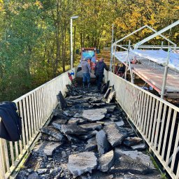 Karol Szwajcer - Fragment zniszczonej nawierzchni mostu z gruzem i ekipą remontową w tle, widok na jesienny las i pomarańczowy ciągnik.
