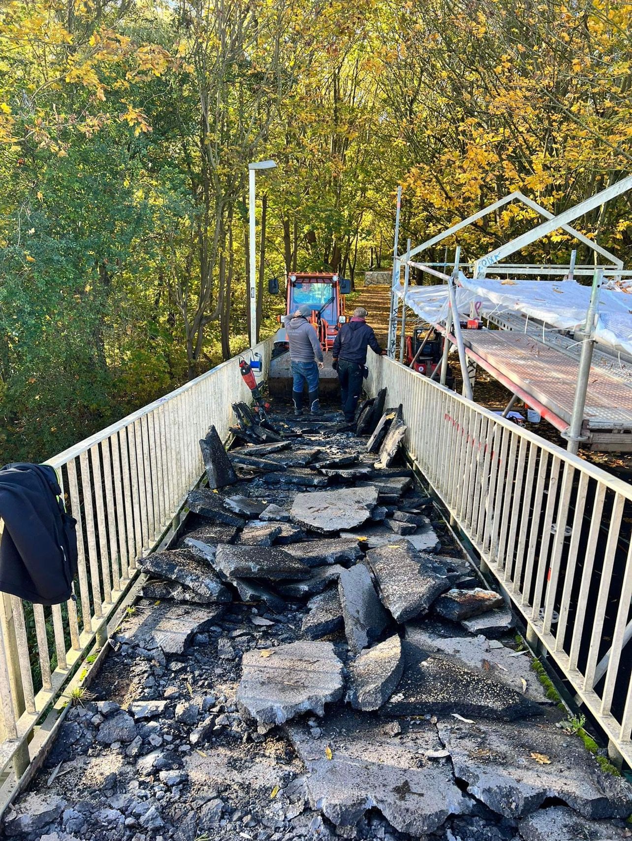 Fragment zniszczonej nawierzchni mostu z gruzem i ekipą remontową w tle, widok na jesienny las i pomarańczowy ciągnik.