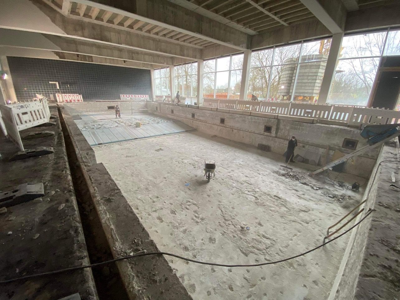 Wnętrze basenu w trakcie prac remontowych, widoczne częściowo usunięte płytki, robotnik zamiatający gruz, w oddali pracownicy i rusztowania zabezpieczające.