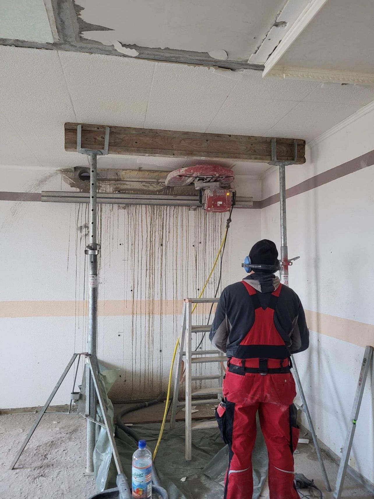 Pracownik w czerwonym kombinezonie, tyłem, obsługuje maszynę do cięcia betonu zamontowaną pod sufitem, wspartą na metalowych podporach. Ściany i sufit są częściowo uszkodzone, widoczne ślady po...