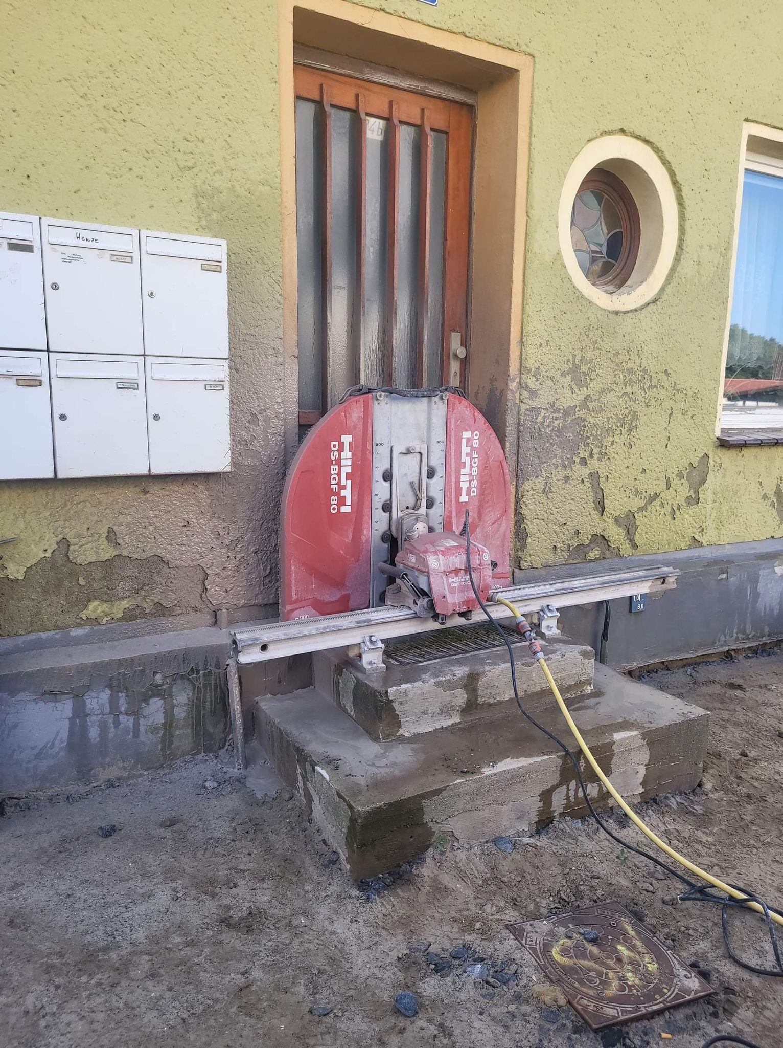 Czerwona piła Hilti DS-BGF 80 przy wejściu do budynku z zieloną elewacją, widoczne schody wejściowe z betonu i fragment okna. Przewody zasilające na ziemi.