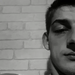 Czarno-biały portret młodego mężczyzny z bliska, z fragmentem białej ściany z cegieł w tle. Ujęcie z prawej strony, z widoczną częścią twarzy i ramienia.