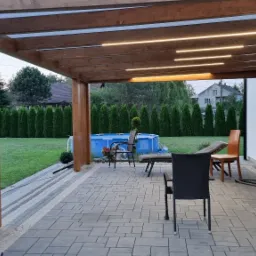 Zadaszony taras z drewnianą konstrukcją, oświetleniem LED i widokiem na basen w ogrodzie.