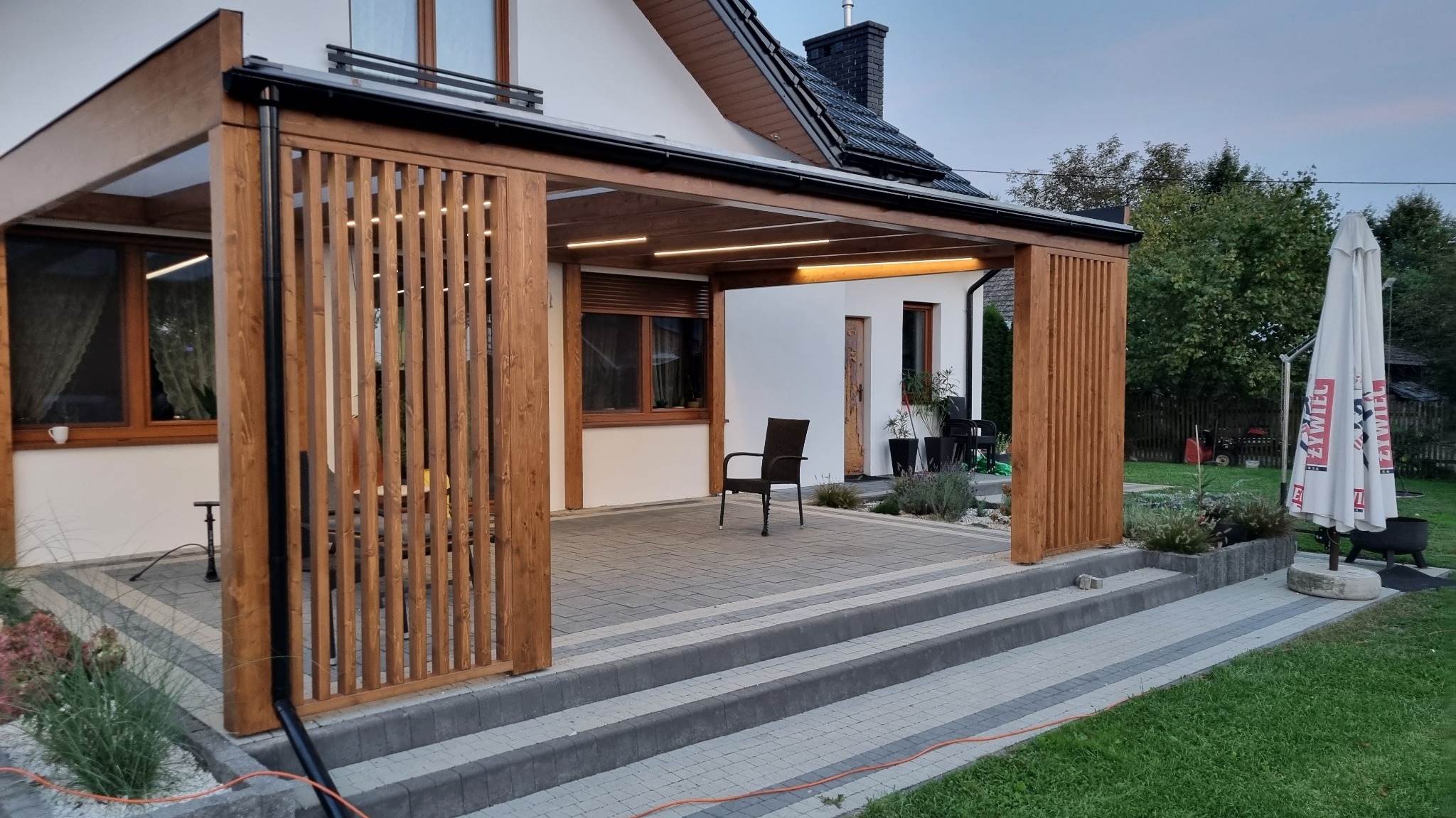 Drewniana pergola z pionowymi lamelami, oświetleniem LED i betonowym tarasem, widok z zewnątrz na dom z ogrodem.