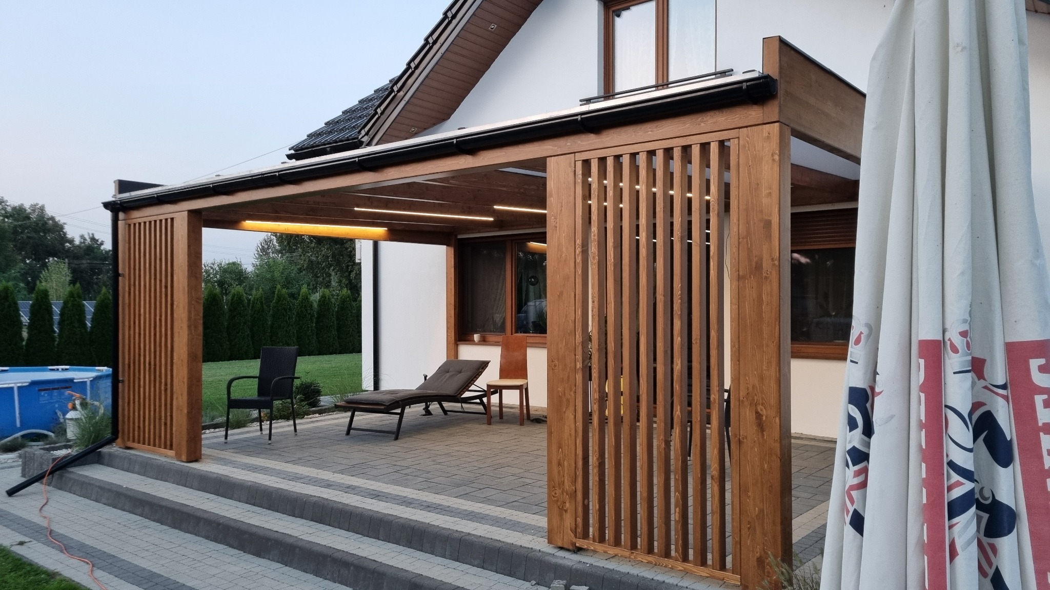 Zadaszona konstrukcja z drewna z oświetleniem LED, na tarasie z szarej kostki, z widocznymi meblami ogrodowymi i fragmentem basenu w tle.