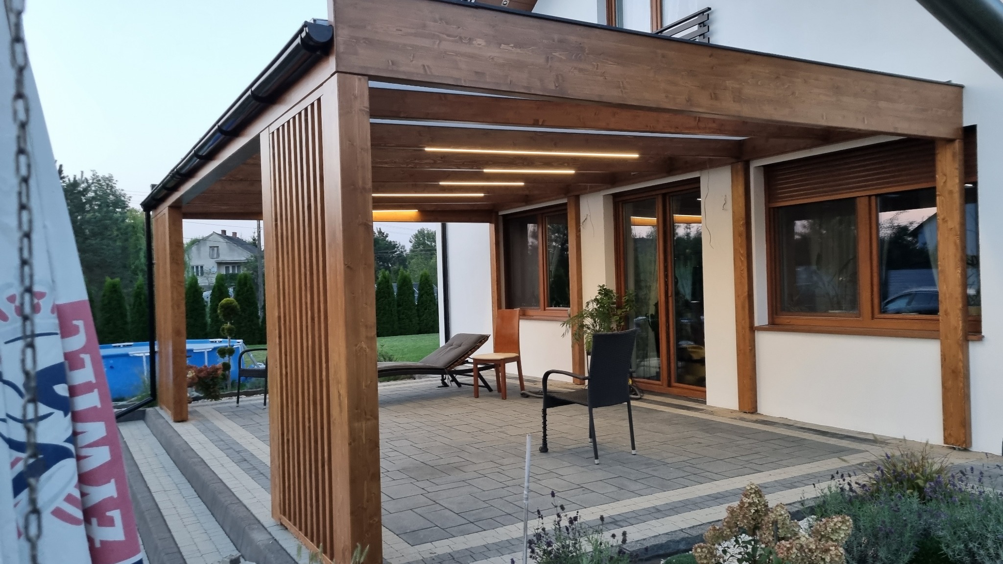 Zadaszony taras z drewnianą konstrukcją i subtelnym oświetleniem LED, widok na ogród z basenem i dom w tle.