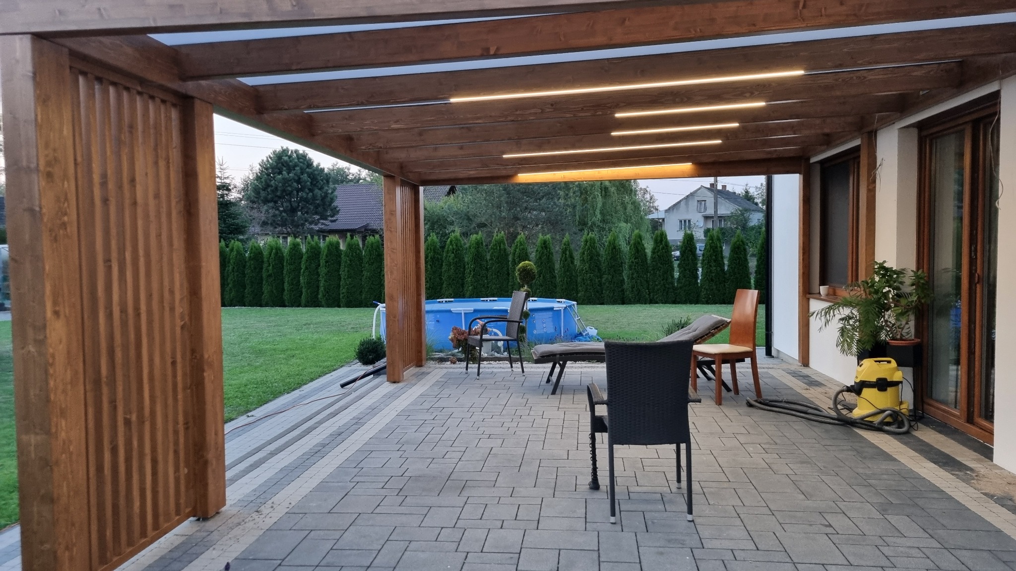 Zadaszony taras z drewnianą konstrukcją, oświetleniem LED i widokiem na basen w ogrodzie.