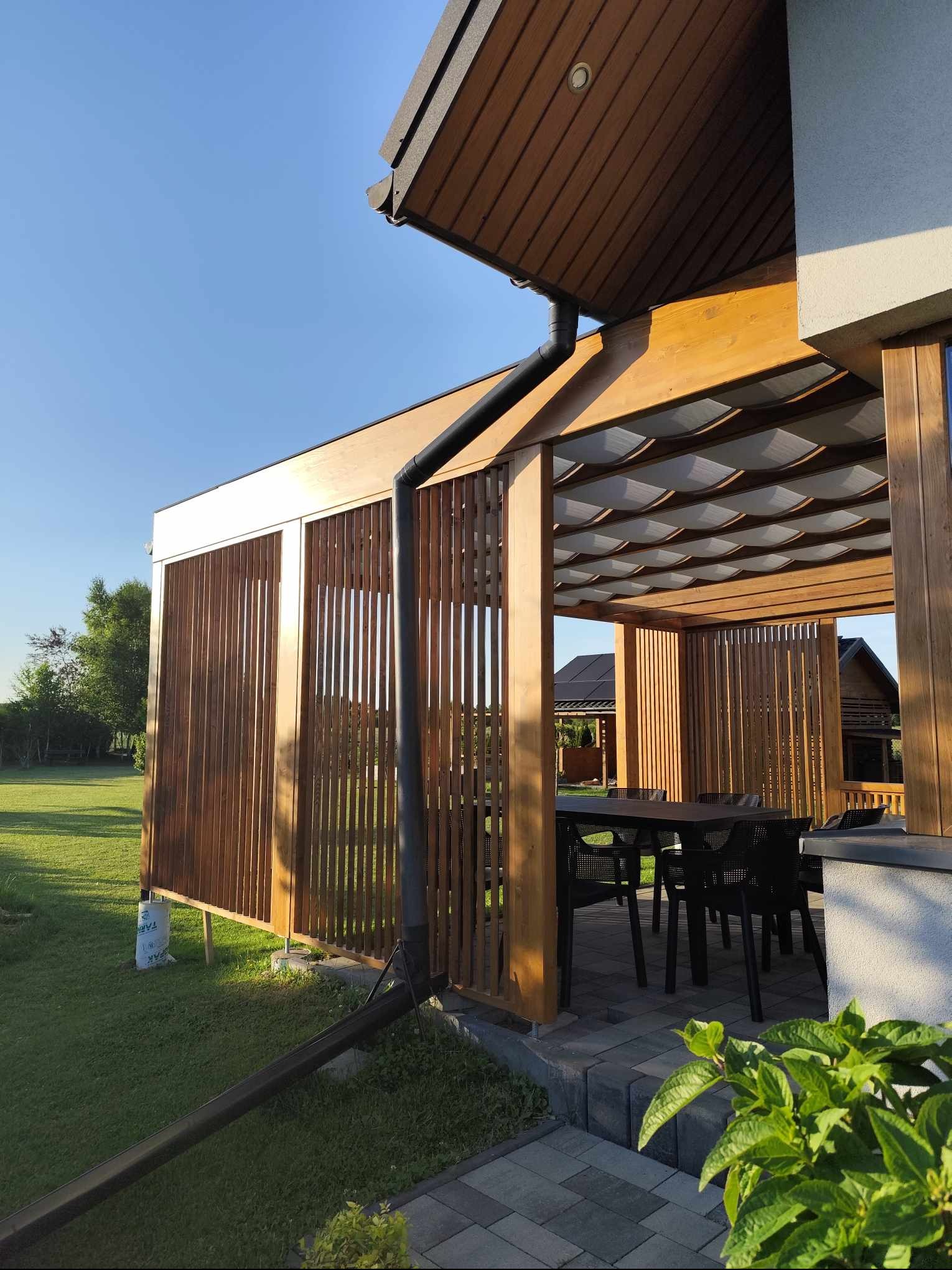 Drewniana konstrukcja z ażurowymi ścianami i zadaszeniem z tkaniny, wyposażona w stół i krzesła, na tle zadbanego trawnika i zabudowań.