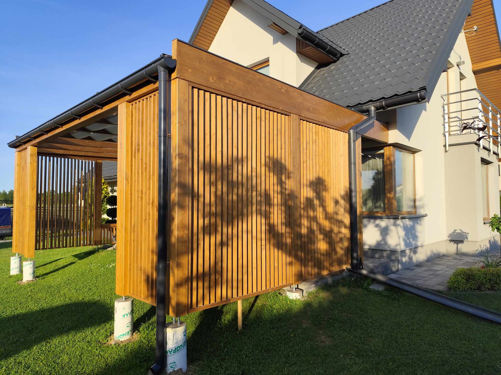 Drewniana konstrukcja z pionowych listew, prawdopodobnie element większej całości, wsparta na betonowych fundamentach, z widocznym systemem rynnowym i cieniem drzewa na ścianie, na tle domu...