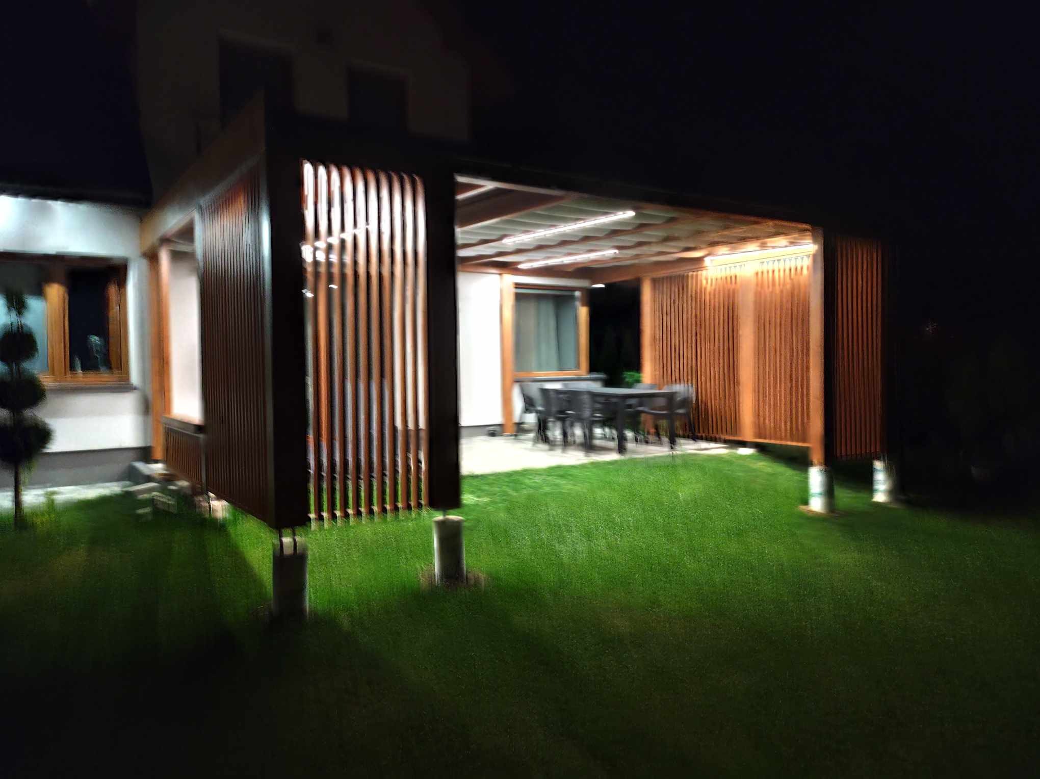 Oświetlona drewniana pergola z lamelami i zadaszeniem tkaninowym, widoczna nocą z trawnika, z betonowymi podstawami słupów.