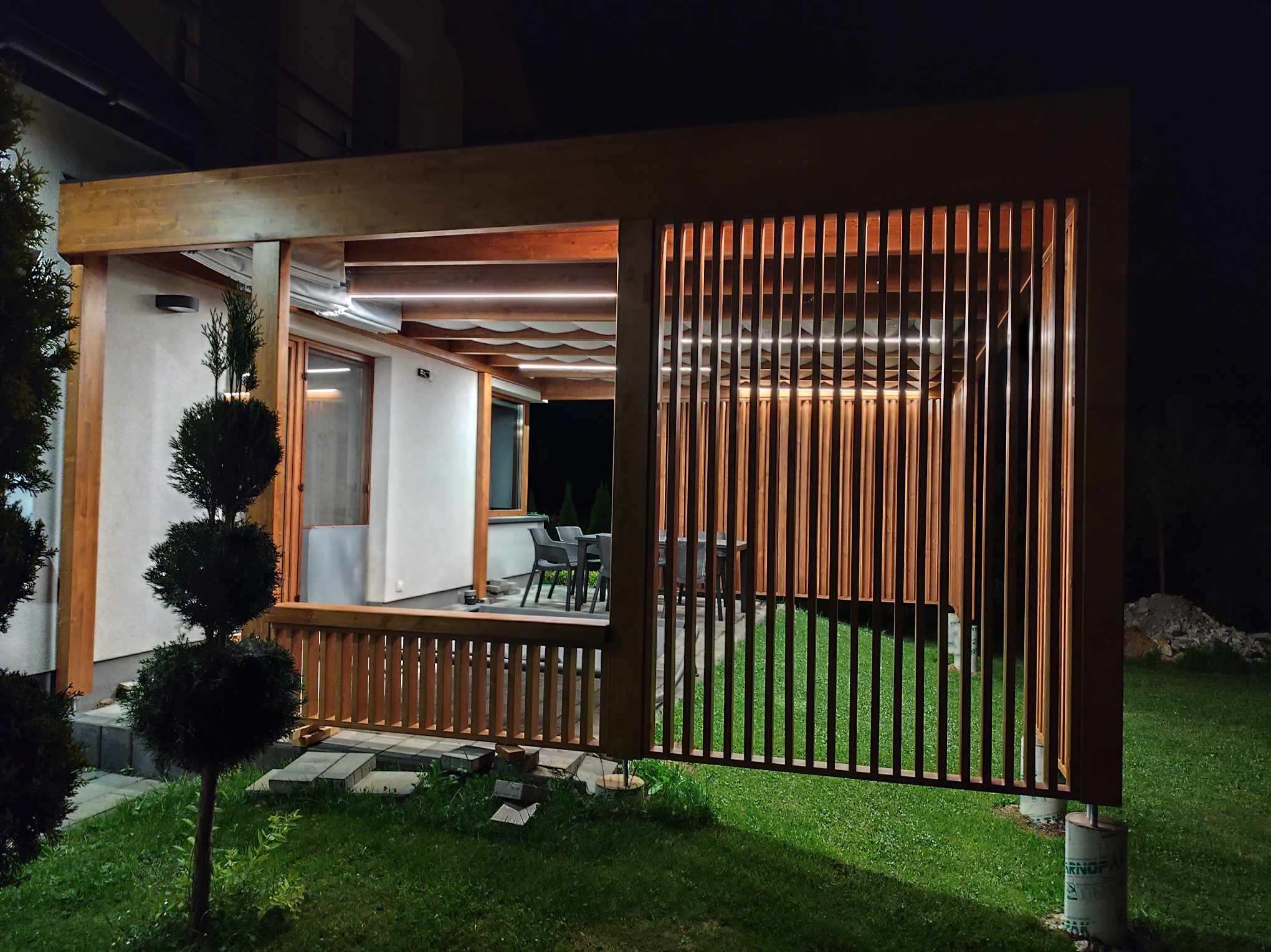 Nowoczesna drewniana pergola z pionowymi lamelami oświetlona od wewnątrz, widok z zewnątrz na tle nocnego ogrodu z trawnikiem i formowanymi krzewami.