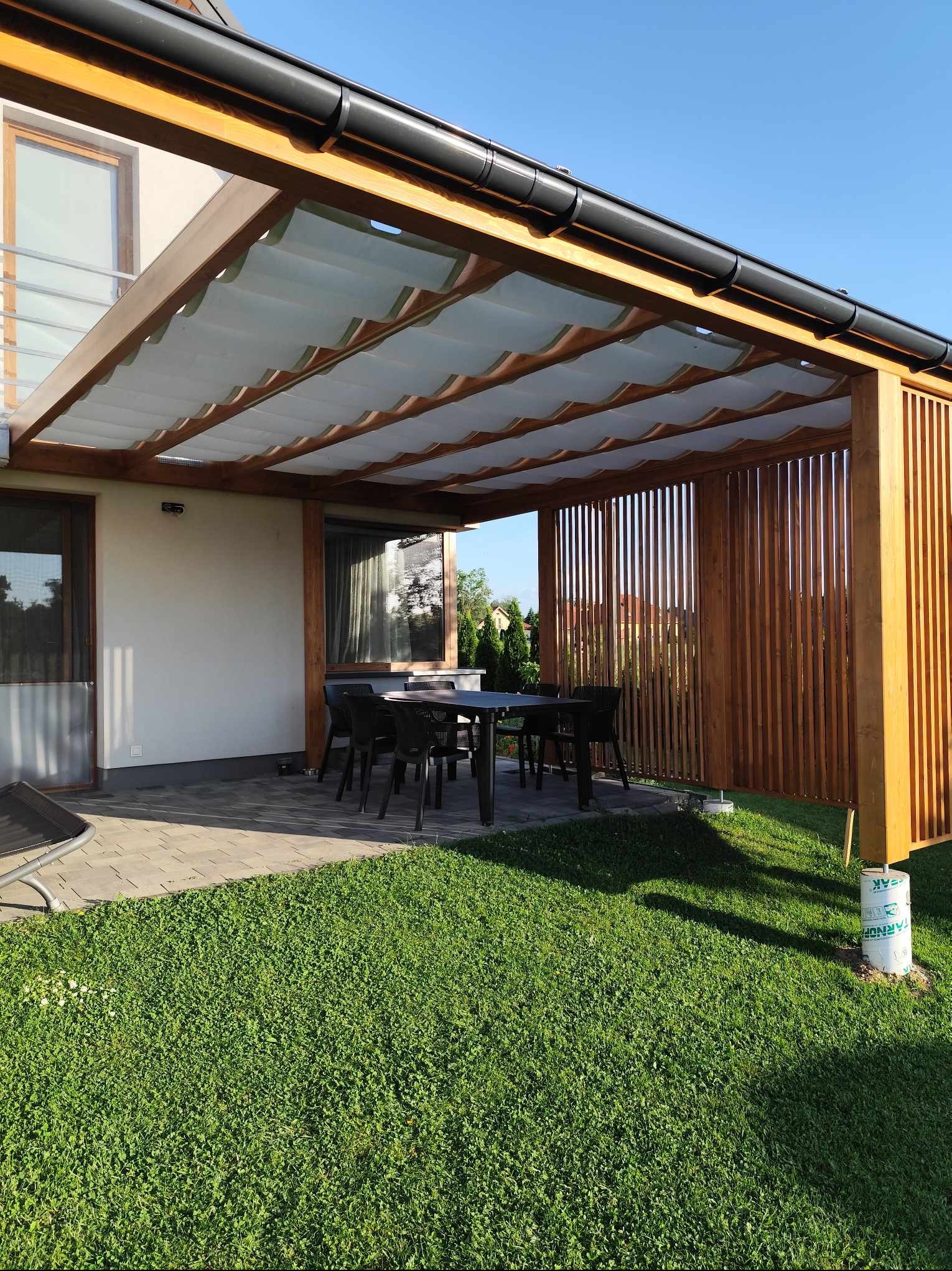 Drewniana konstrukcja pergoli z rozsuwanym materiałowym zadaszeniem, osłaniająca taras z czarnym stołem i krzesłami, widok z trawnika w słoneczny dzień.