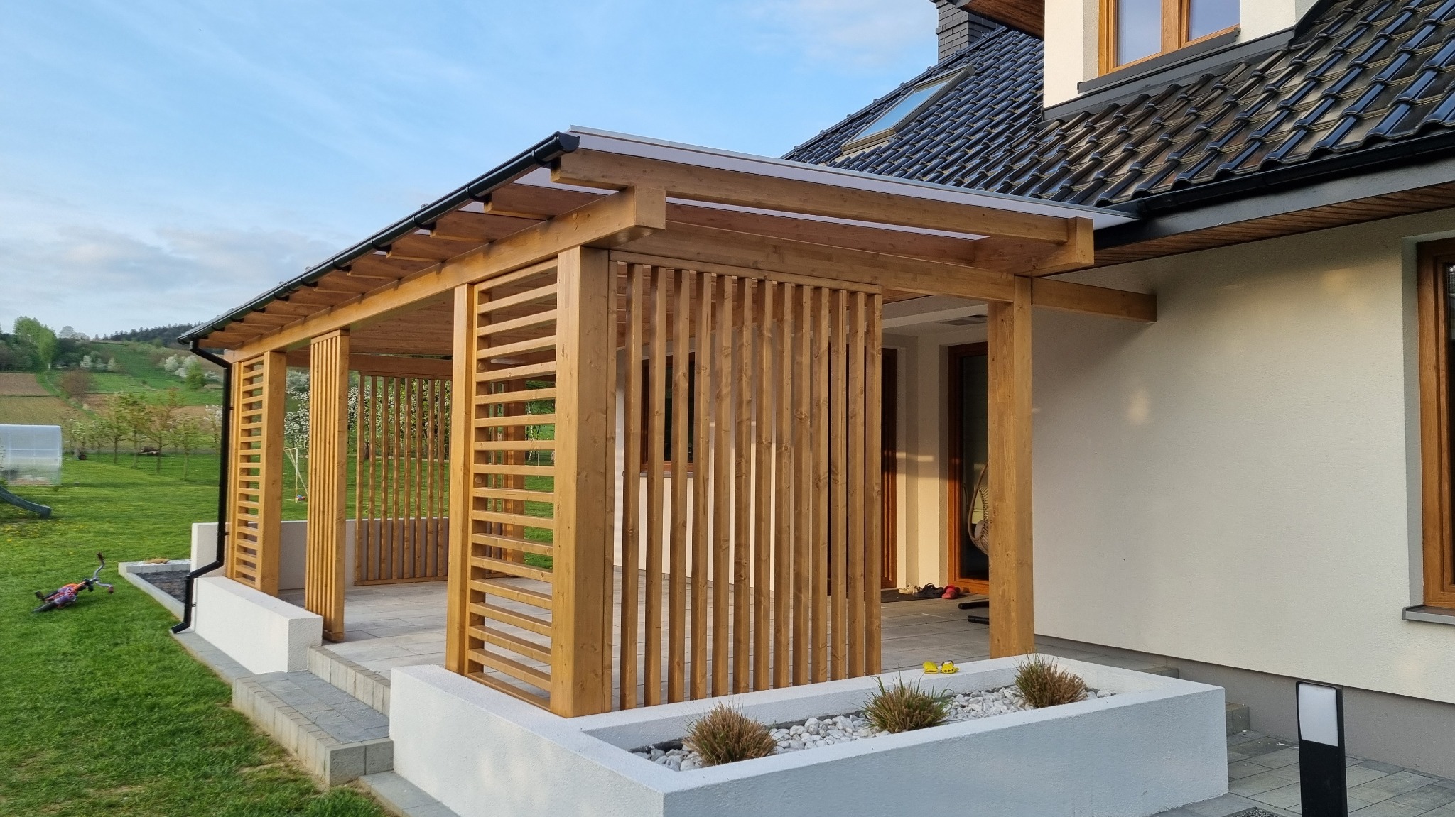 Drewniana pergola z lamelami i zadaszeniem z poliwęglanu przy nowoczesnym domu, widok z zewnątrz, w tle zielony krajobraz.