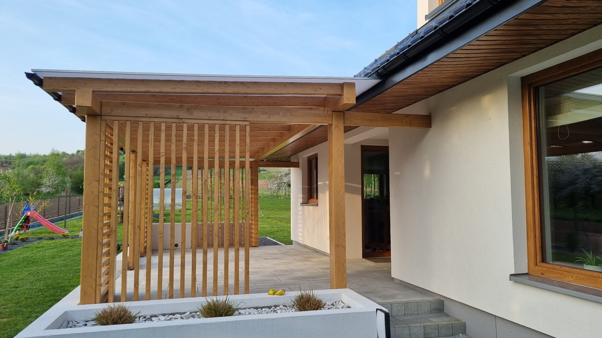 Drewniana konstrukcja z pionowymi listwami przylegająca do domu, z widocznym fragmentem tarasu z szarych płyt, w tle zielony trawnik i zjeżdżalnia.