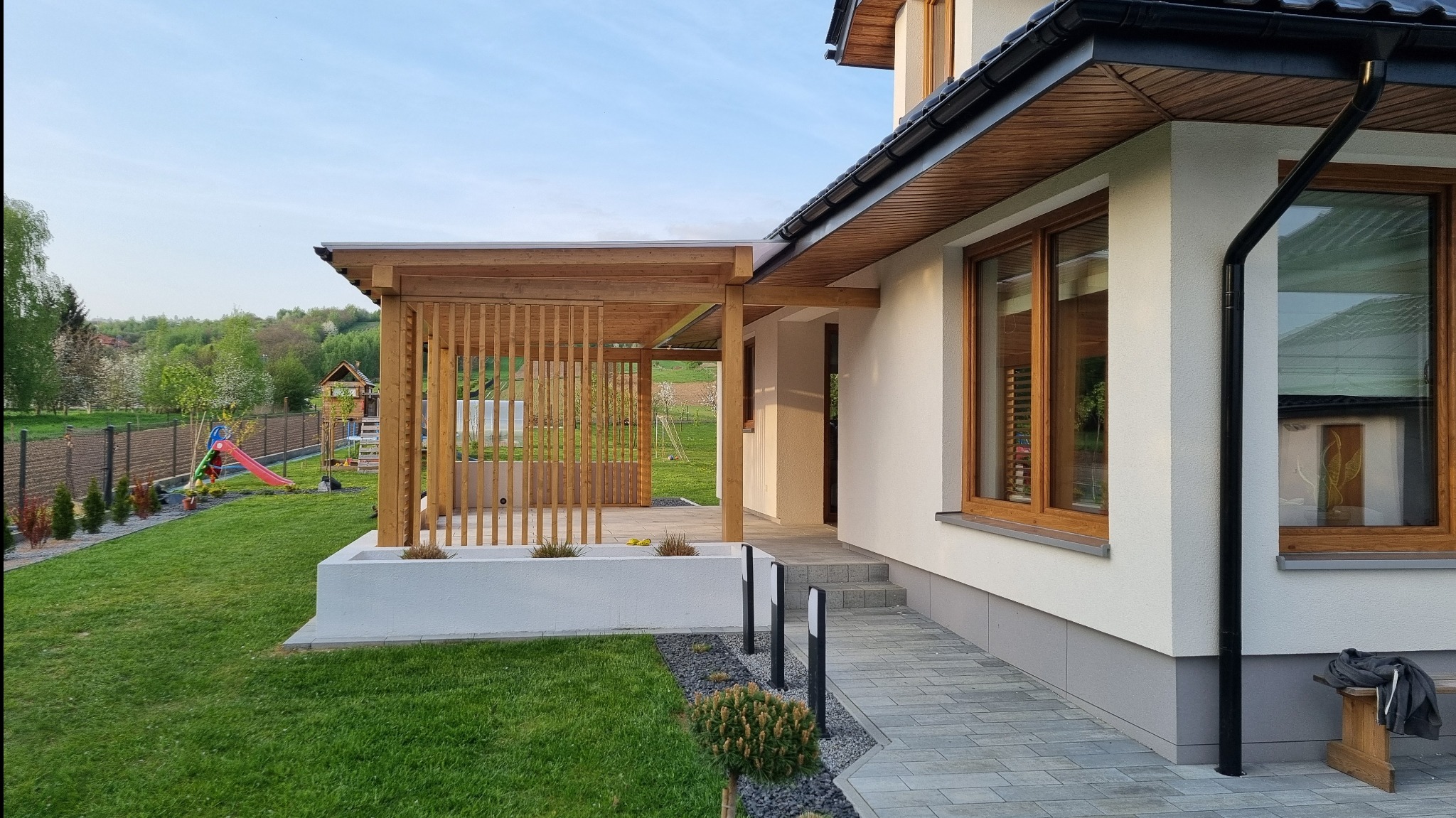 Nowoczesna drewniana pergola z pionowymi listwami, przylegająca do białego domu z drewnianymi oknami, widok na zadbany trawnik i plac zabaw w tle.
