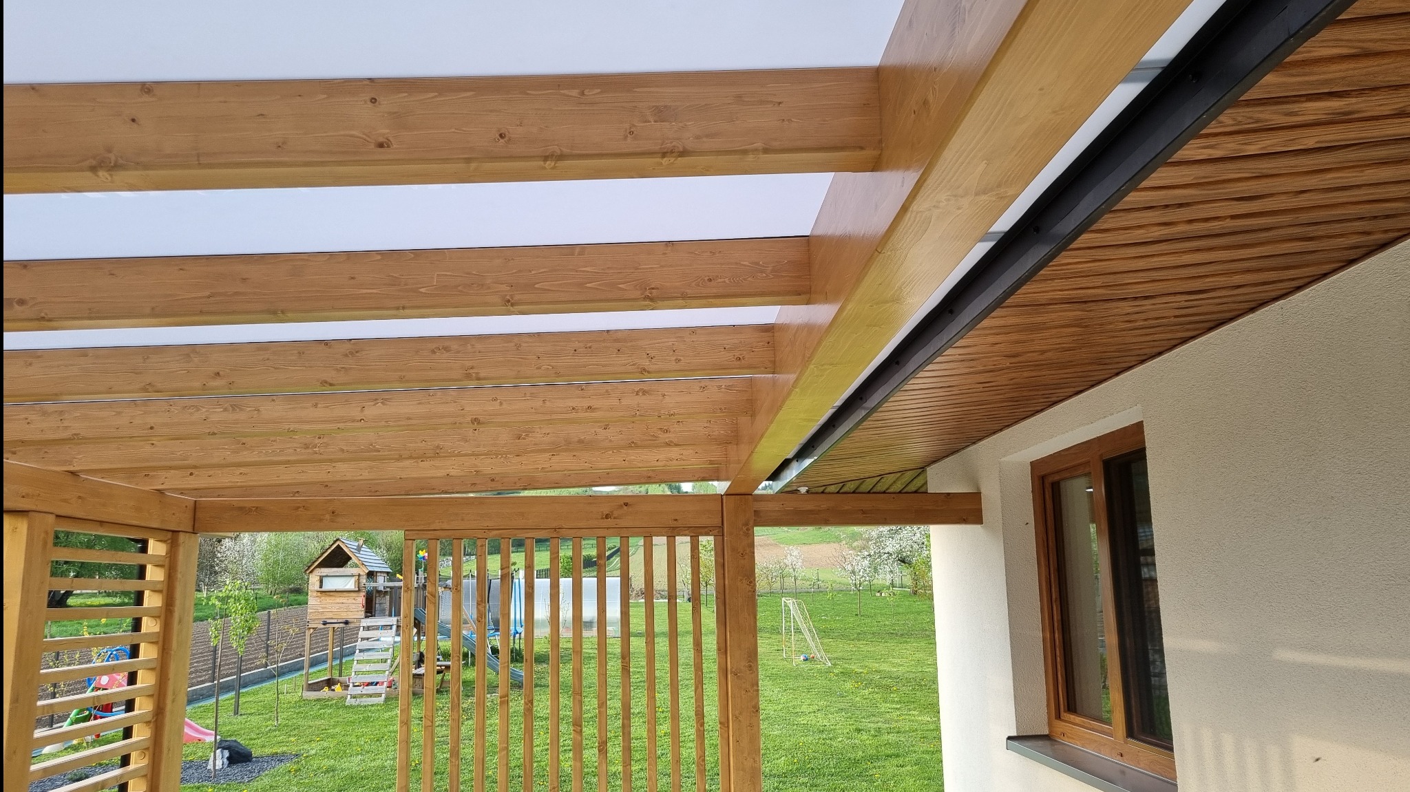 Drewniana pergola z przezroczystym zadaszeniem, widok od wewnątrz na zielony ogród z domkiem dla dzieci i trampoliną, z elementami czarnej metalowej konstrukcji.