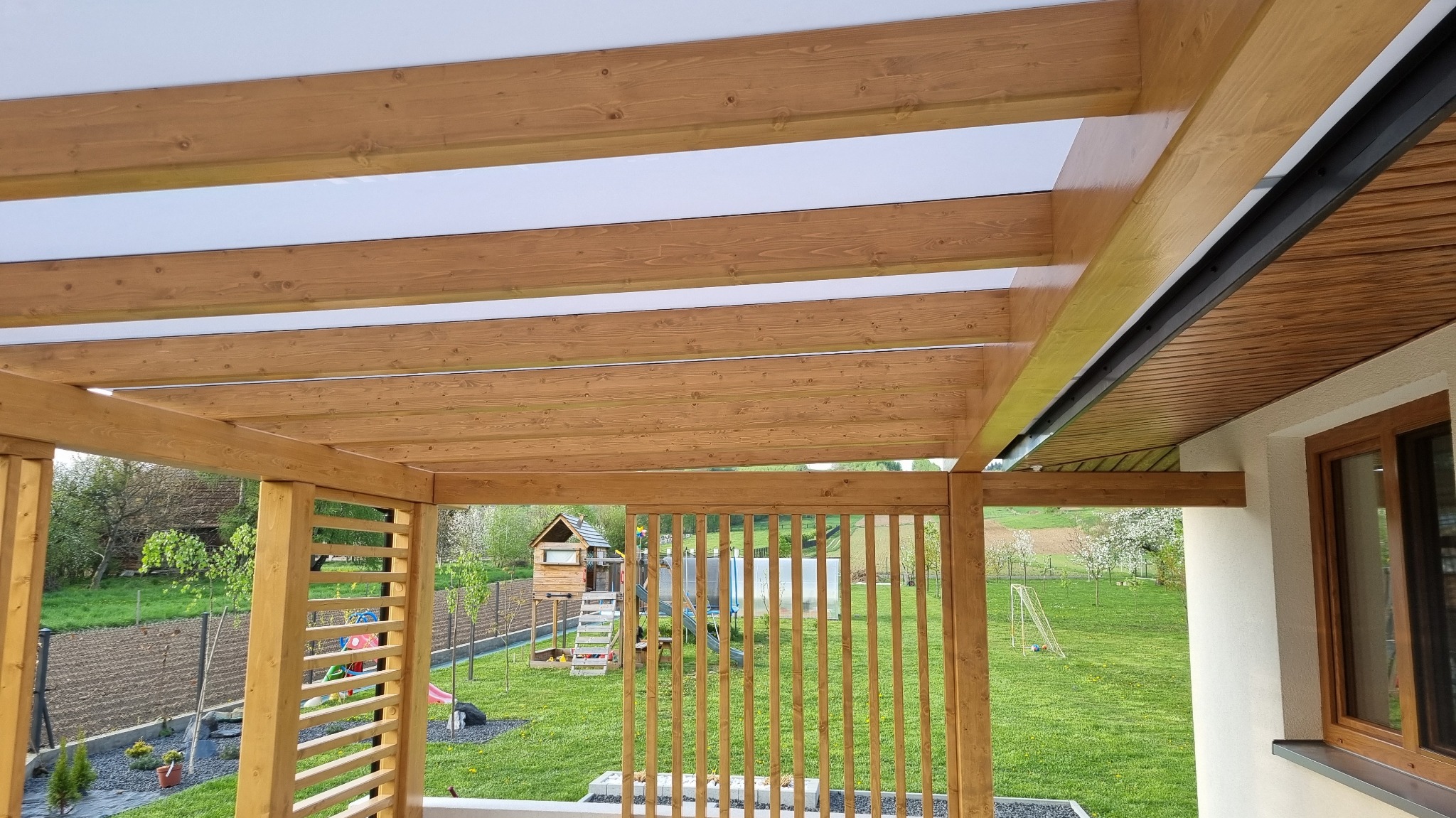 Zadaszenie tarasu z drewnianą konstrukcją i białym, przepuszczającym światło dachem, widok na zielony ogród z domkiem dla dzieci w tle.