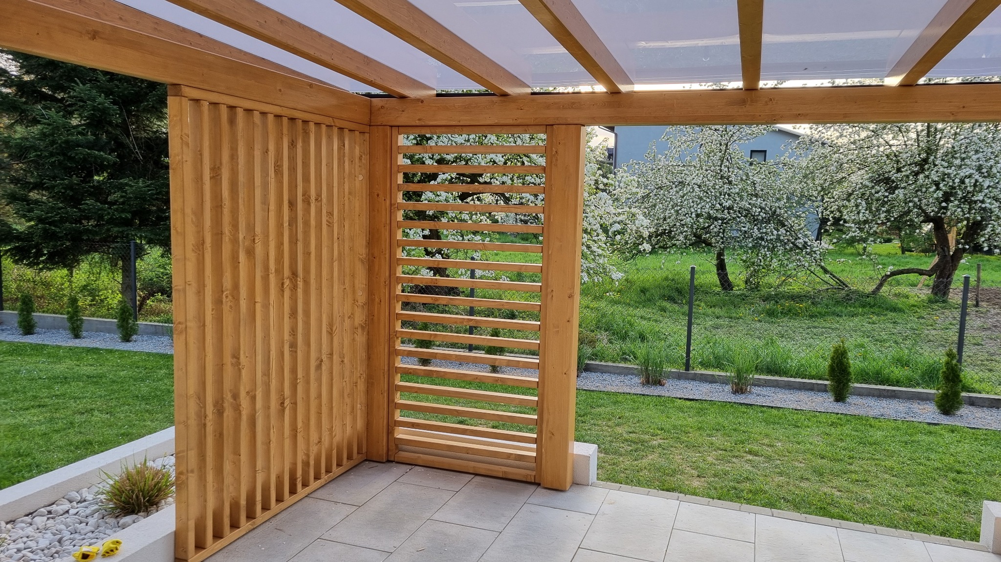 Drewniana konstrukcja z ażurową ścianką i przezroczystym dachem, widok na zielony ogród z kwitnącymi drzewami