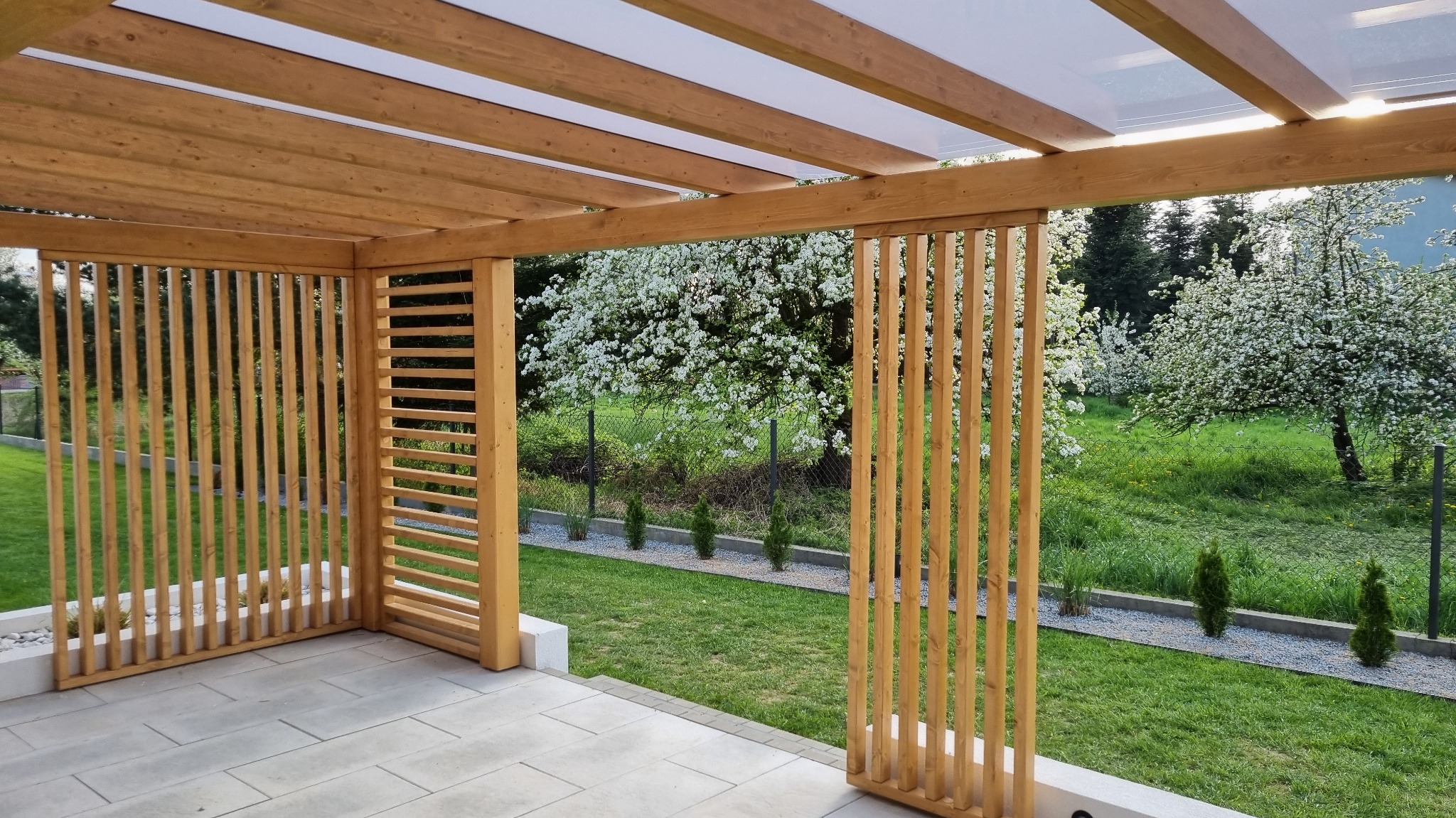 Drewniana konstrukcja z ażurowymi ściankami i przezroczystym zadaszeniem, widok z wnętrza na kwitnące drzewa w ogrodzie.