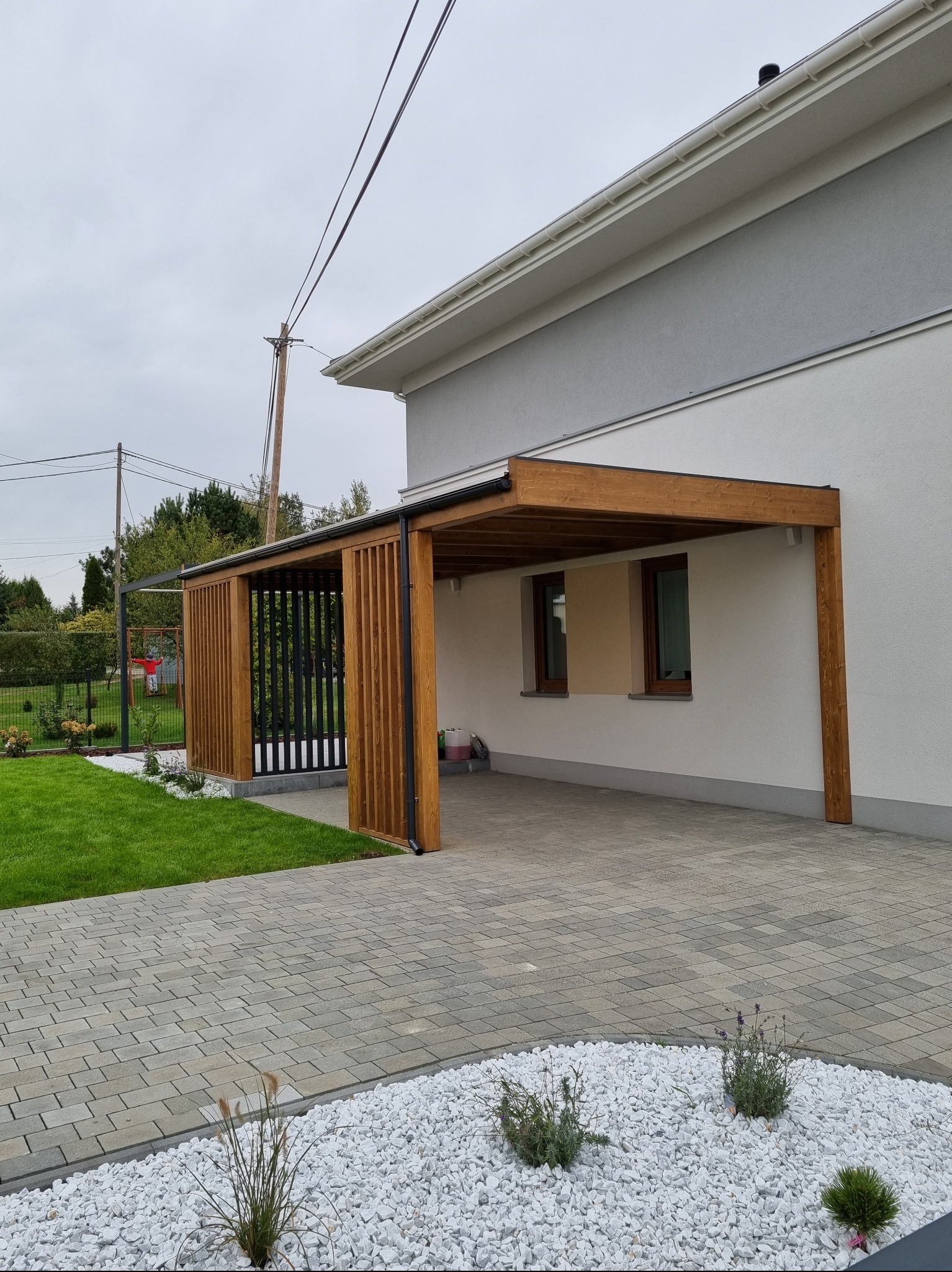 Nowoczesna pergola z drewnianymi elementami i czarnymi pionowymi panelami, przylegająca do jasnego domu z szarym akcentem, otoczona trawnikiem i ścieżką z kostki brukowej z białym kamieniem ozdobnym.