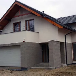 Nowoczesny dom jednorodzinny z szarą cegłą klinkierową na parterze, białym tynkiem na piętrze, balkonem z metalową balustradą i bramą garażową segmentową.