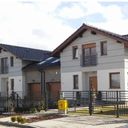 Dwa bliźniacze domy z szarą elewacją, brązowymi ramami okiennymi i czarnym dachem, otoczone niskim, czarnym ogrodzeniem panelowym z furtką i żółtą skrzynką pocztową.