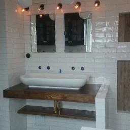 Biała, owalna umywalka nablatowa z trzema niebieskimi kurkami zamontowana na drewnianym blacie z półką, otoczona białymi, prostokątnymi płytkami ściennymi, podwieszona pod dwoma lustrami...