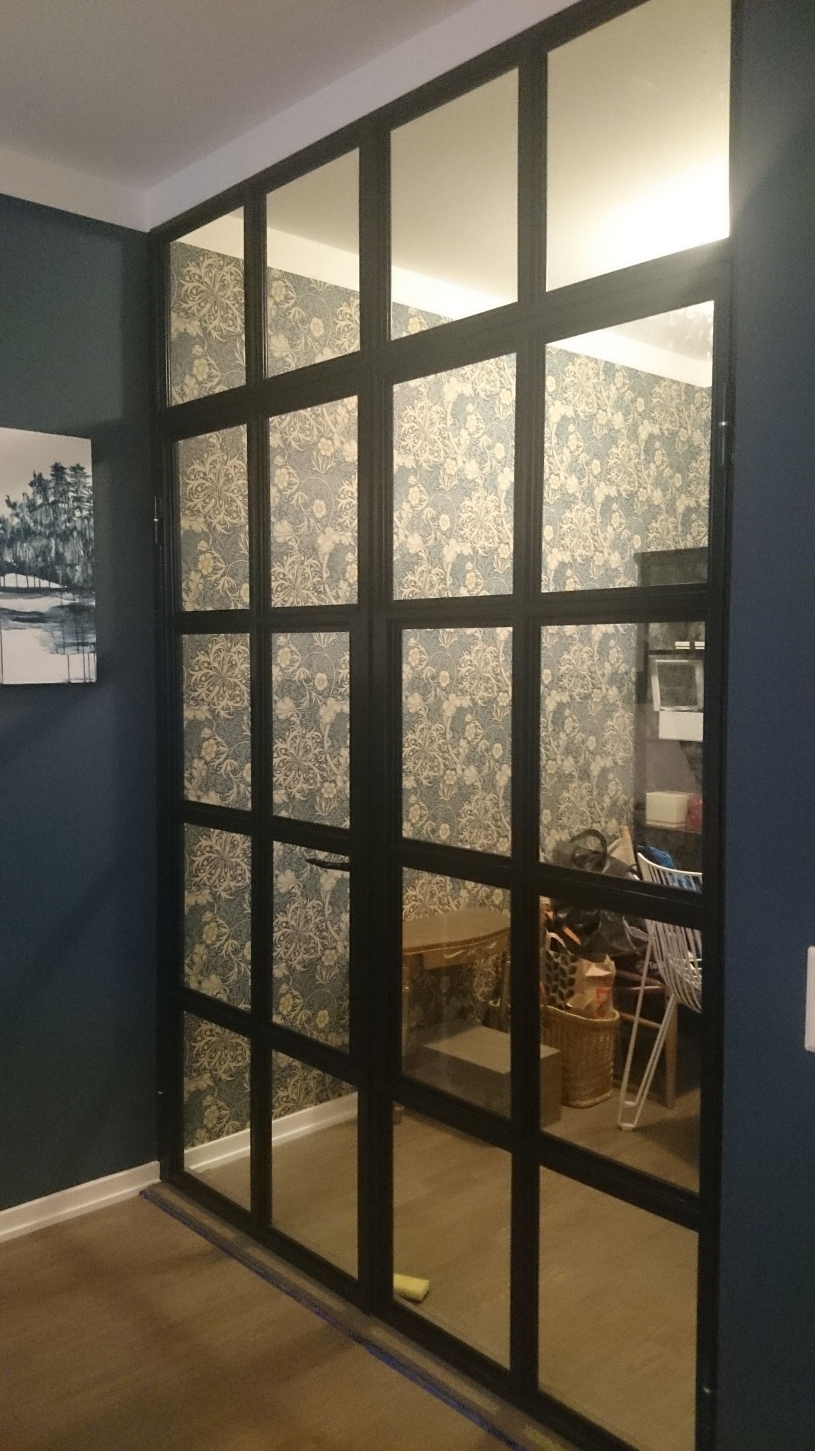 Przeszklone drzwi przesuwne z czarnymi szprosami, oddzielające pomieszczenie z wzorzystą tapetą, widoczne elementy wystroju wnętrza, takie jak obraz na ścianie i meble w pokoju obok.