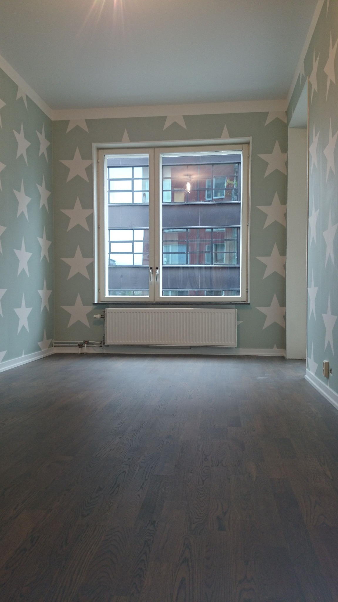 Pusty pokój z ciemną drewnianą podłogą, ścianami w pastelowym odcieniu z białymi gwiazdami i oknem z widokiem na budynek.