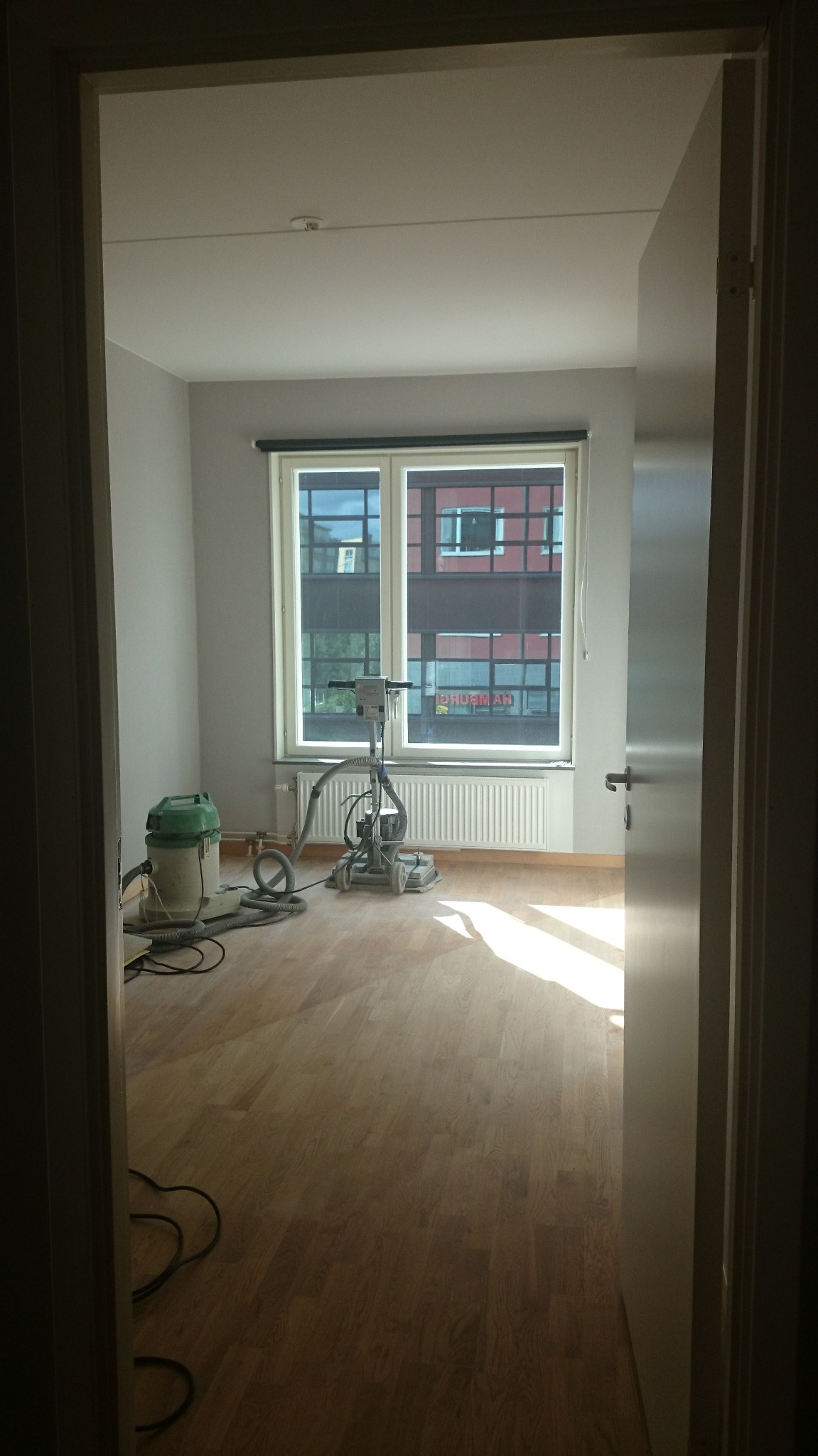 Pokój w trakcie renowacji podłogi drewnianej, widoczna szlifierka do parkietu, odkurzacz przemysłowy i promienie słońca wpadające przez okno.