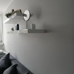 Świeżo pomalowana szara ściana z dwoma białymi półkami, na których stoją dekoracje: roślina, lustro i świeca, widok z dołu na szarą sofę.