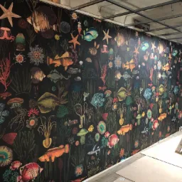 Ściana pokryta czarną tapetą z motywem morskiego życia: ryby, rozgwiazdy, meduzy i rośliny, widoczne także fragmenty sufitu i drabina malarska.