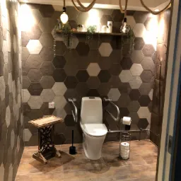 Nowoczesna toaleta z heksagonalnymi płytkami na ścianach, dekoracyjnymi żarówkami zwisającymi na linach, drewnianą półką z roślinami i ozdobami, oraz poręczami dla osób z niepełnosprawnościami.