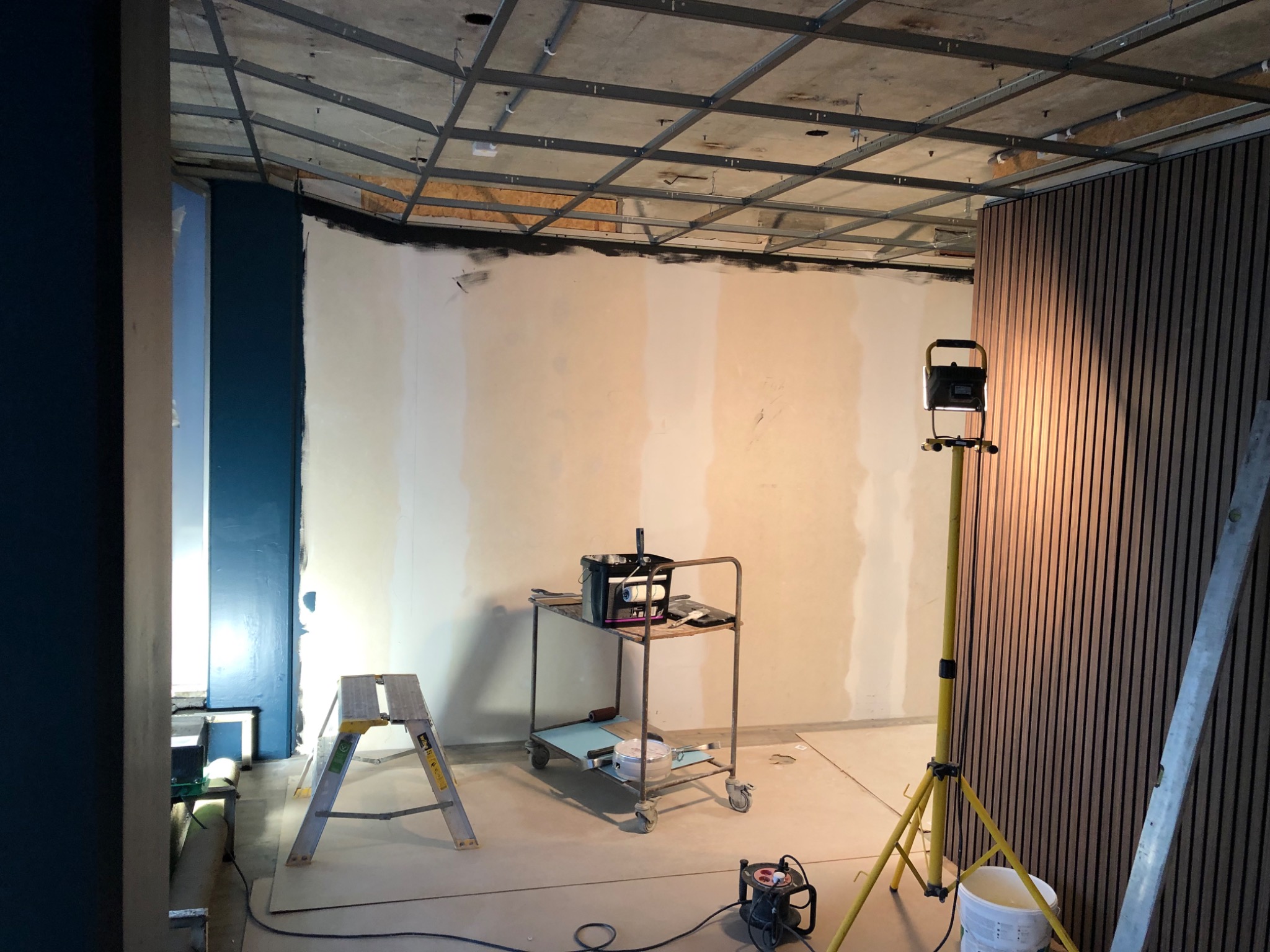 Wnętrze w trakcie prac przygotowawczych: surowy sufit z metalową konstrukcją, ściany częściowo zagruntowane, widoczny wózek z narzędziami malarskimi, drabina i lampa budowlana.