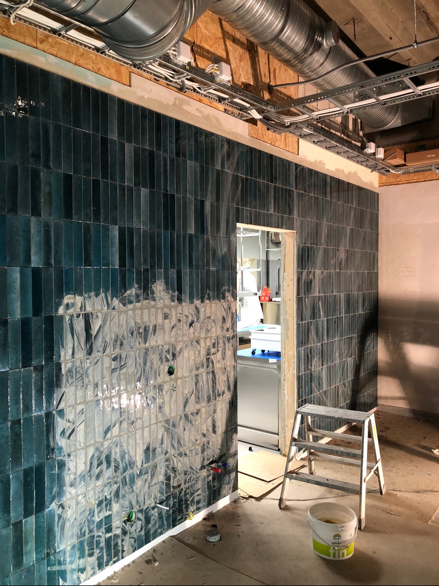 Ściana w trakcie wykańczania z pionowo ułożonych, błyszczących, turkusowych płytek. Widoczne ślady fugowania, drabina i wiadro z zaprawą. Na suficie instalacja wentylacyjna i elektryczna.