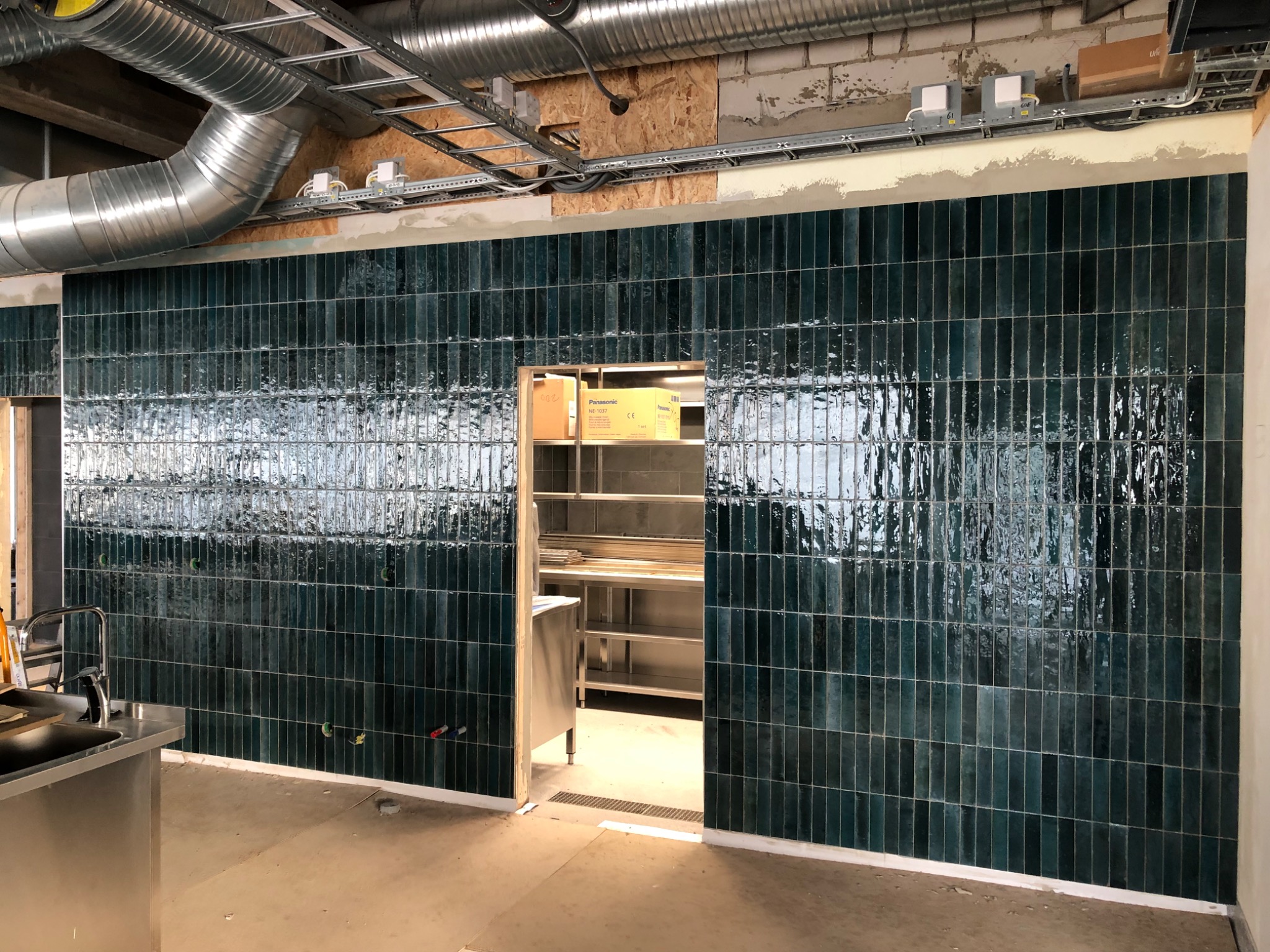 Ściana wyłożona pionowymi, błyszczącymi, ciemnozielonymi płytkami w pomieszczeniu w trakcie prac wykończeniowych, widoczne rury wentylacyjne pod sufitem i metalowe regały w przejściu