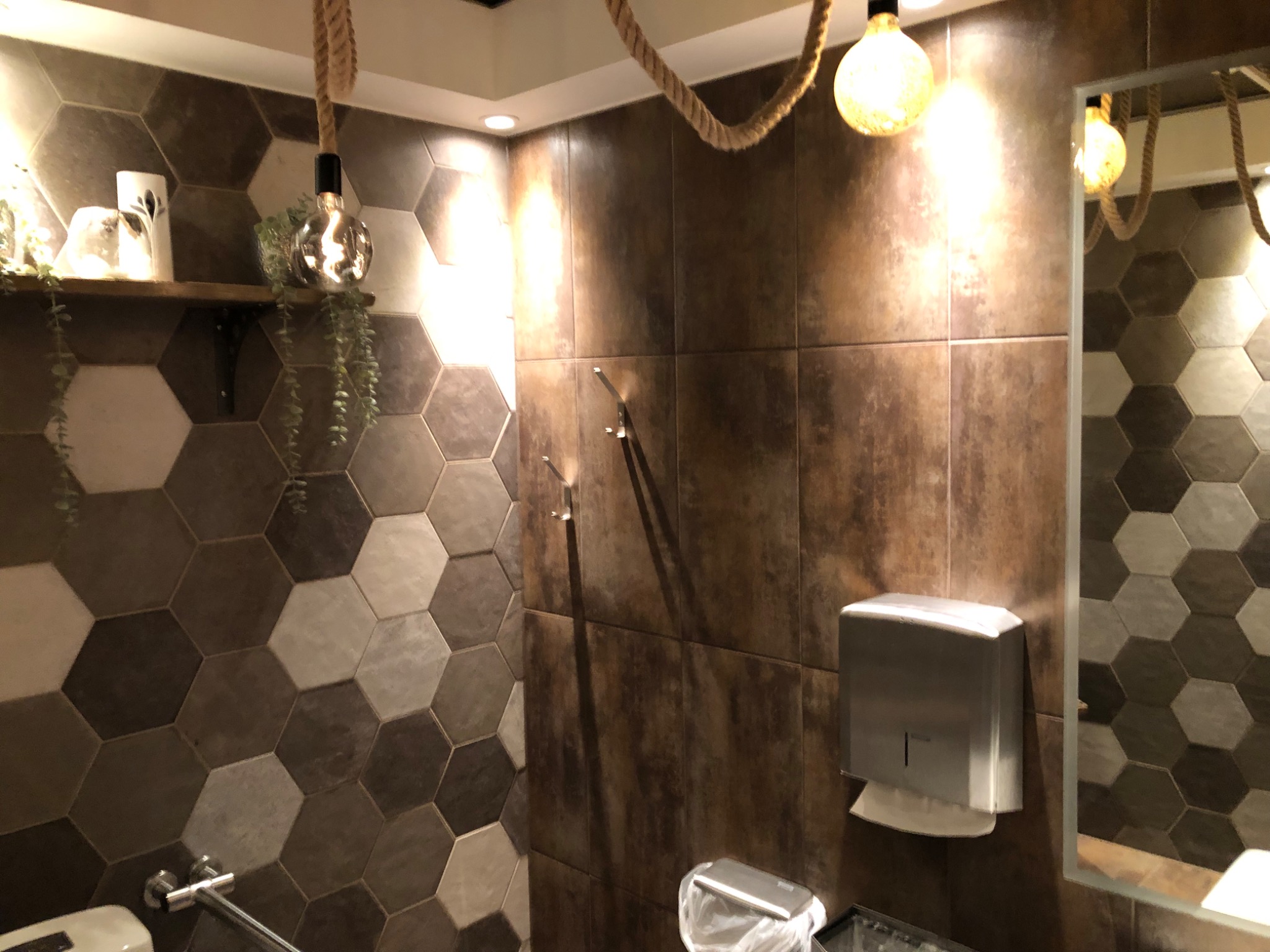 Nowoczesna łazienka z heksagonalnymi płytkami w odcieniach szarości i brązu, oświetlona żarówkami na sznurze, z widocznym dozownikiem ręczników papierowych i fragmentem toalety.