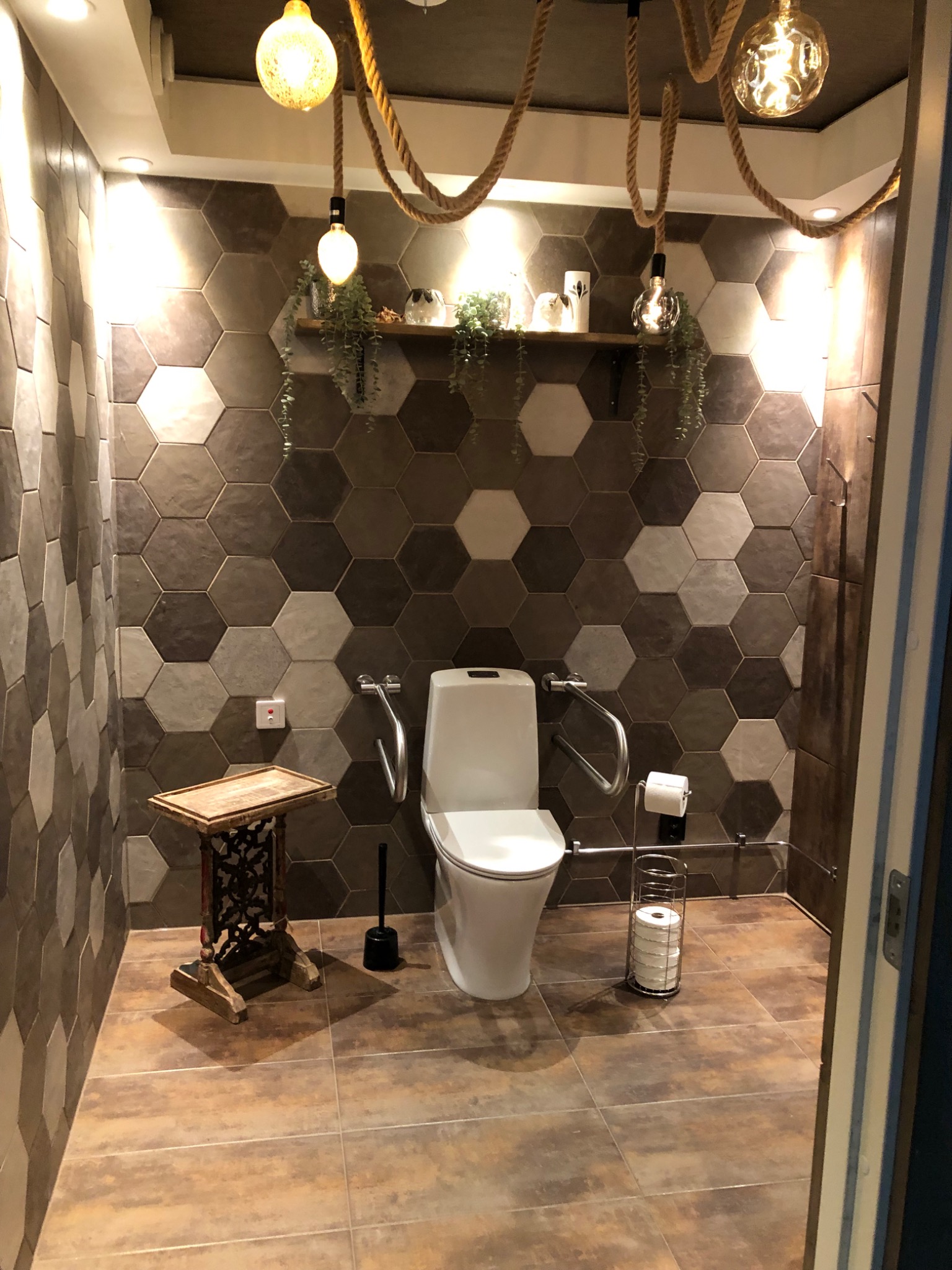 Nowoczesna toaleta z heksagonalnymi płytkami na ścianach, dekoracyjnymi żarówkami zwisającymi na linach, drewnianą półką z roślinami i ozdobami, oraz poręczami dla osób z niepełnosprawnościami.
