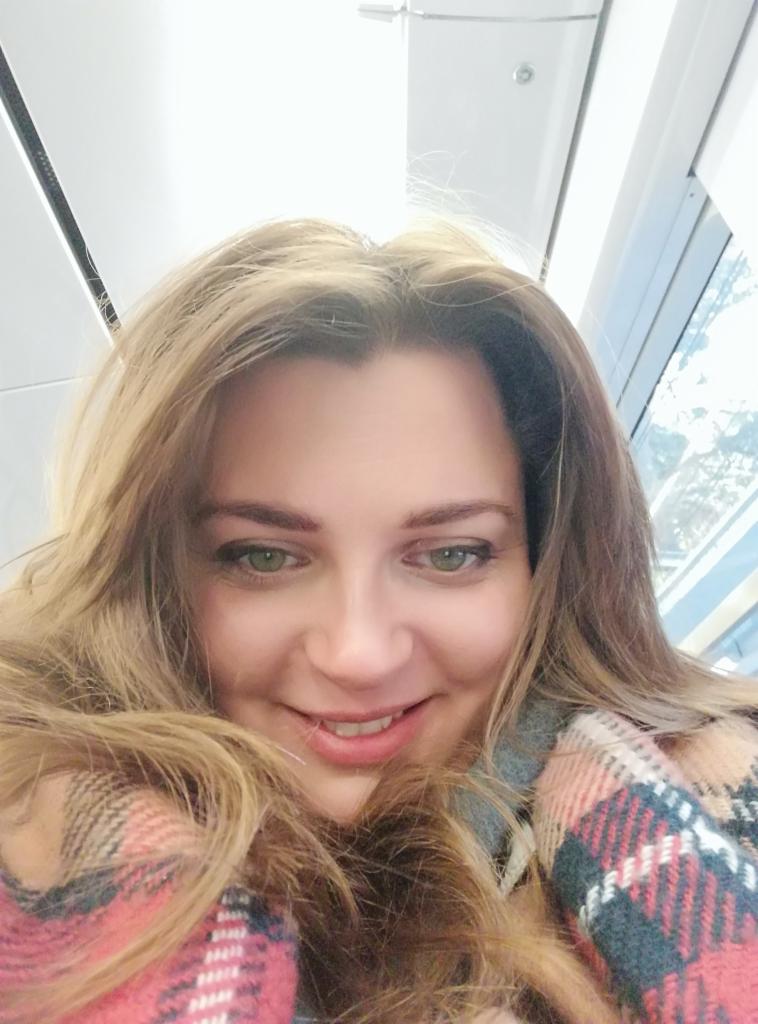 Uśmiechnięta kobieta z długimi blond włosami, ubrana w kraciasty szal, spogląda w dół, z widocznym oknem w tle. Jasne, naturalne oświetlenie.