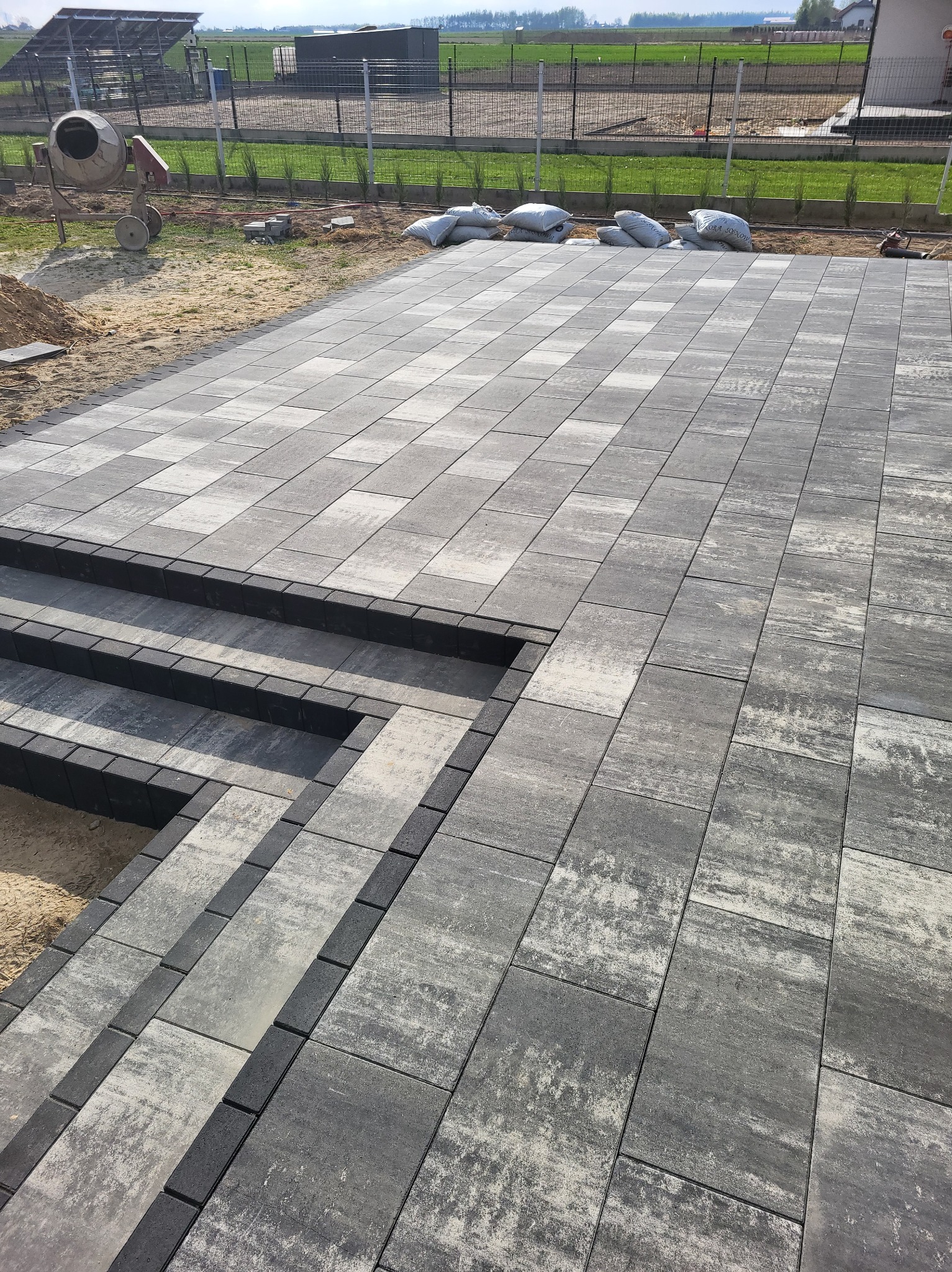 Ułożona kostka brukowa w odcieniach szarości tworząca taras ze schodami. W tle widoczna betoniarka i worki z materiałem, sugerujące trwające prace wykończeniowe.