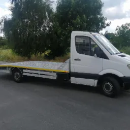 Biała laweta Mercedes z żółtymi akcentami, zaparkowana w Zgierzu na tle zieleni. Widok z boku, lekko z dołu, podkreślający długość platformy.