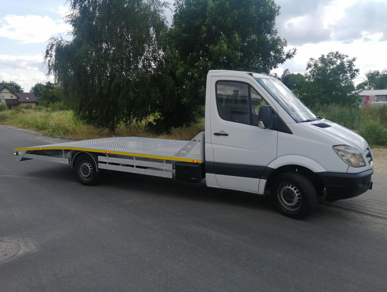 Biała laweta Mercedes z żółtymi akcentami, zaparkowana w Zgierzu na tle zieleni. Widok z boku, lekko z dołu, podkreślający długość platformy.